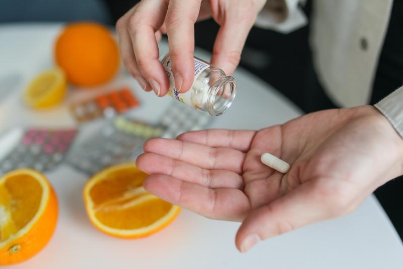 A person pours a pill onto the hand with oranges on the table in the background