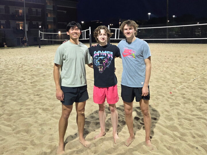 Three people standing on a sand volleyball court at night.