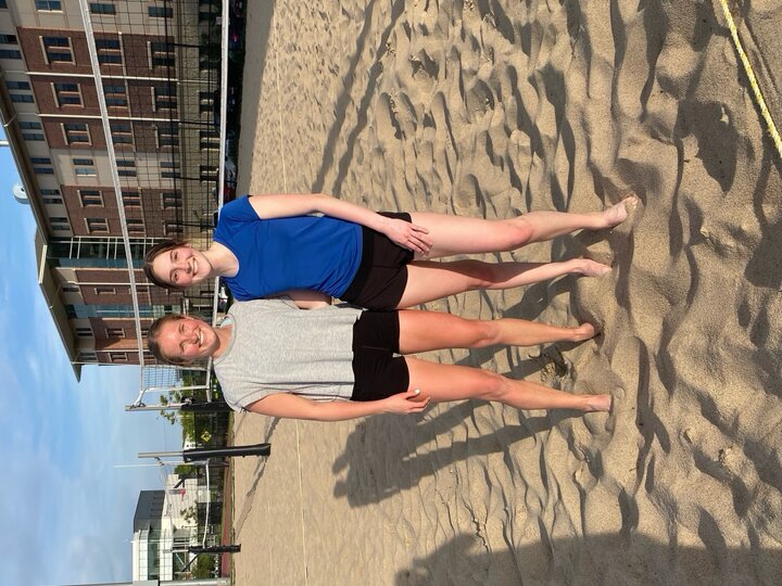 Two people stand on a sandy beach volleyball court near buildings under a blue sky.