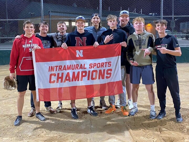 Group of students posing with flag on softball infield dirt.