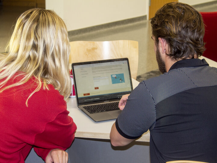 Two people sitting at a table, collaborating while looking at a laptop screen.