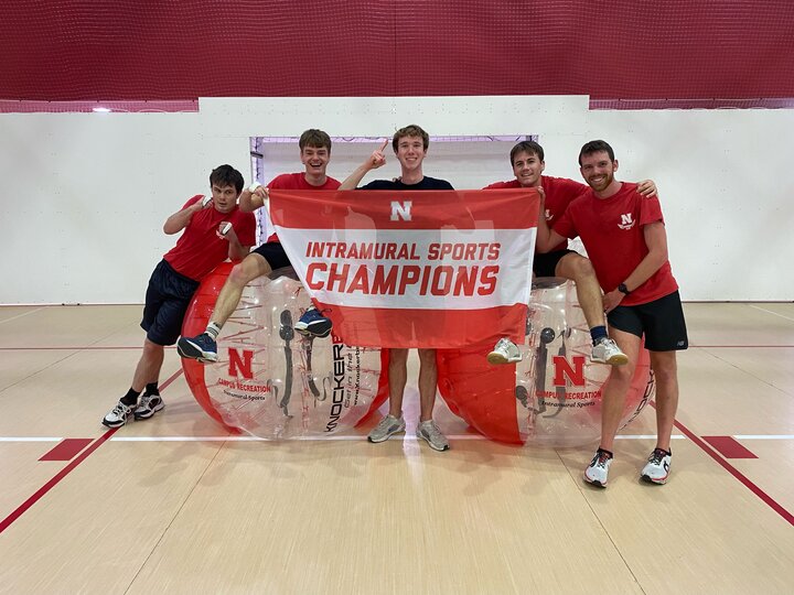 Group holding a banner reading "Intramural Sports Champions" in front of bubble soccer equipment indoors.