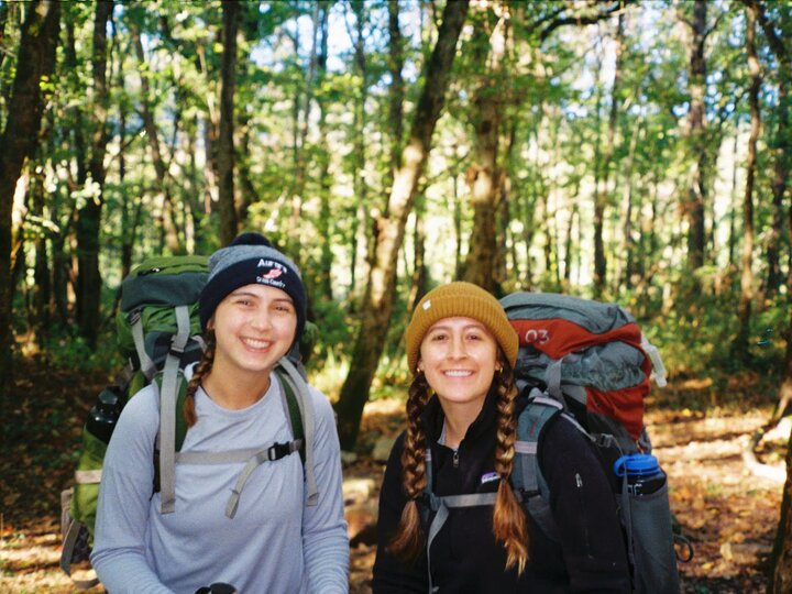 Two women with backpacks smiling in a sunlit forest clearing.