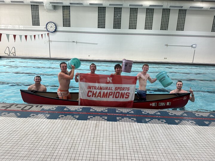 Group of men in a canoe holding buckets and a red "Intramural Sports Champions" banner in a pool.