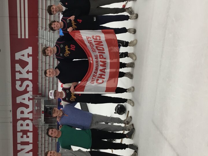 Group of men standing on an ice rink holding a red banner, with "Nebraska" on the wall in the background.