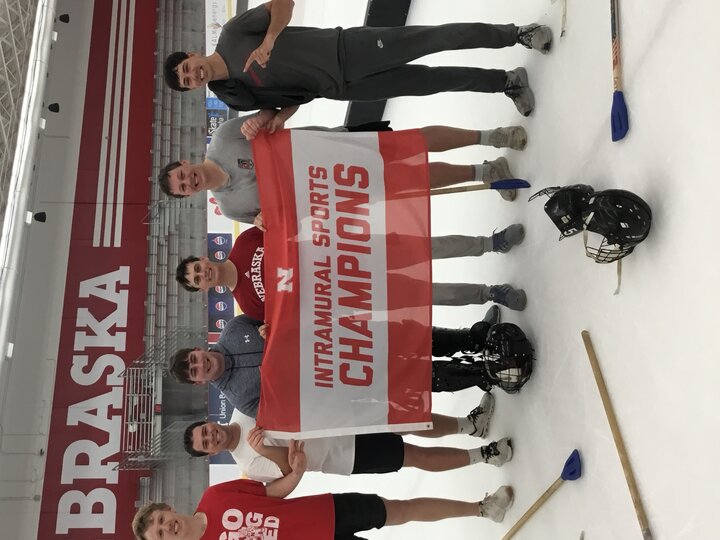 Group of men on an ice rink holding a banner reading "Intramural Sports Champions" with helmets and sticks nearby.