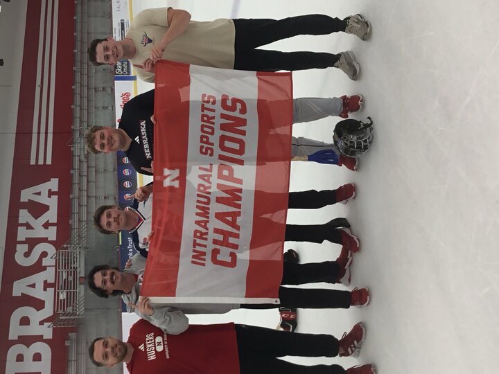 Group of six smiling men on an ice rink holding a red and white banner reading "Intramural Sports Champions."