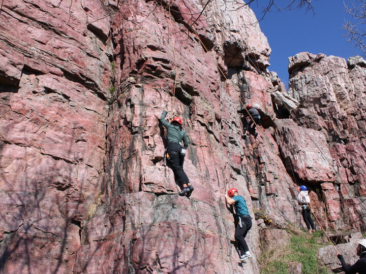 Climbers wearing helmets scale a pink rocky cliff under a clear blue sky.