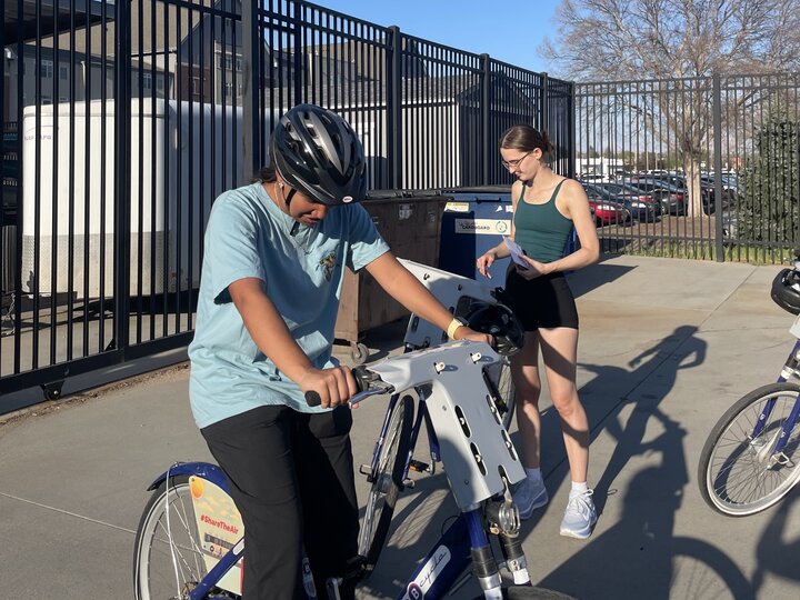A person rides a rental bike while another person stands nearby holding a clipboard outdoors.