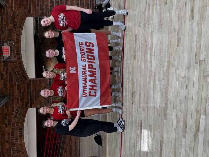 Six women smiling, holding a red banner reading "Intramural Sports Champions" on an indoor court.