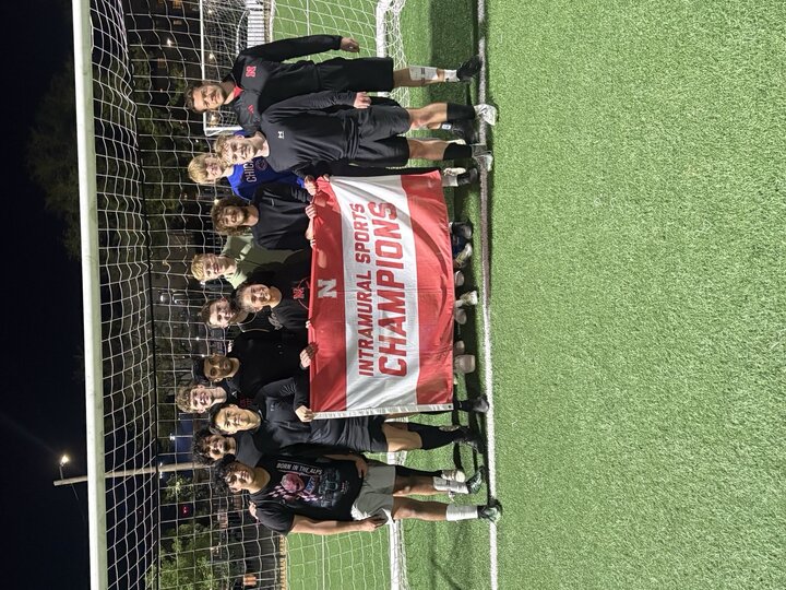 Group of soccer players smiling under a goalpost, holding a red "Intramural Sports Champions" banner.