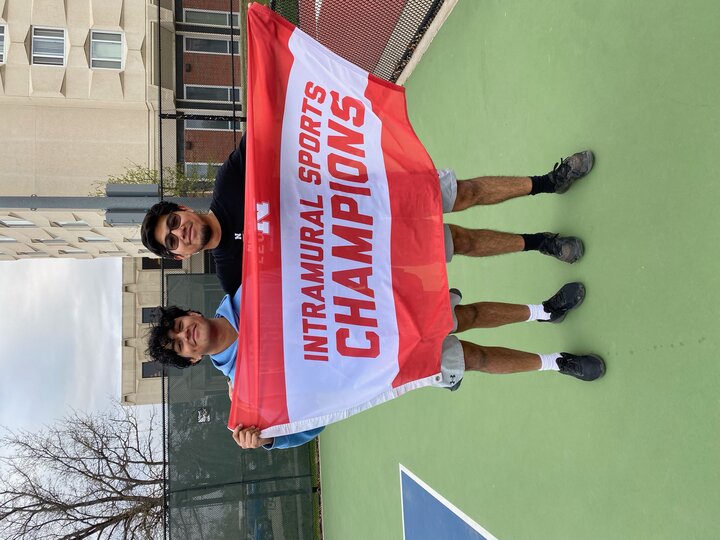 Two people smiling and holding a red and white banner reading "Intramural Sports Champions" on a tennis court.