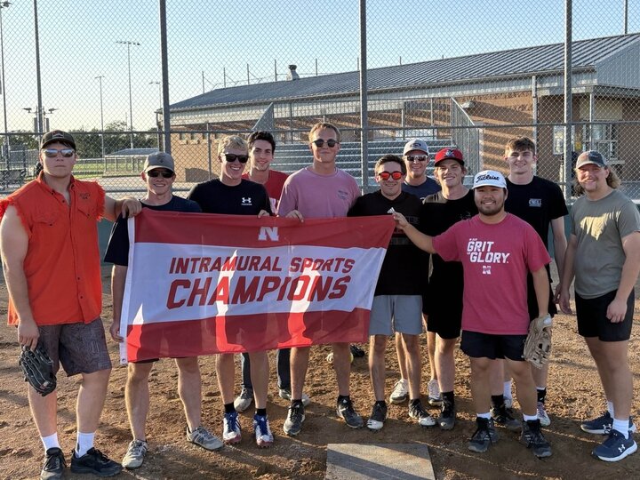 Group of students posing with flag on softball infield dirt.