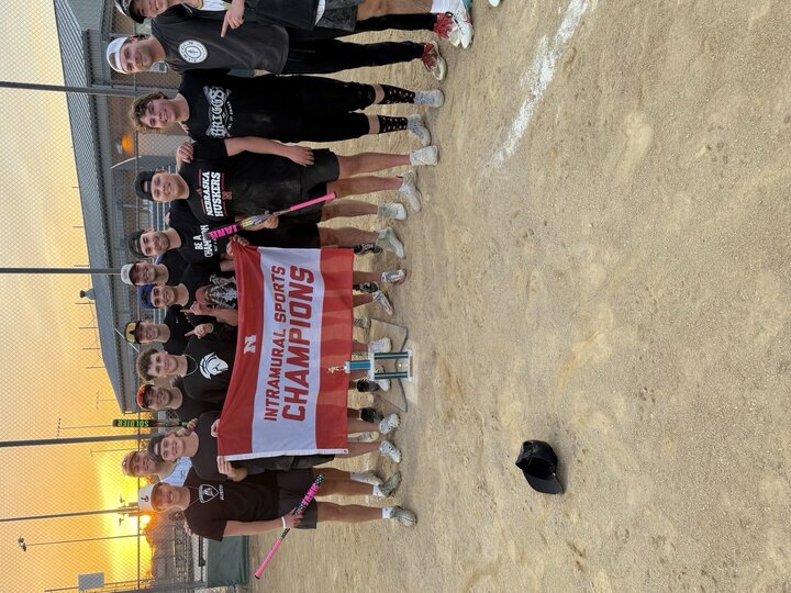 Group holding a "Intramural Sports Champions" banner on a softball field at sunset.