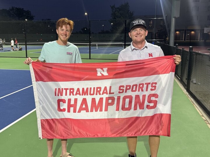 two students posing with flag at tennis courts.