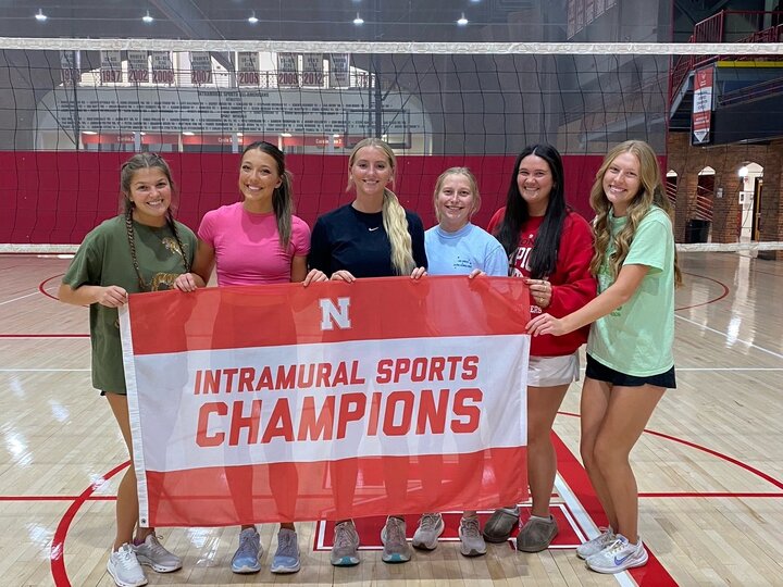 Group of six people holding a red and white "Intramural Sports Champions" banner on a volleyball court.