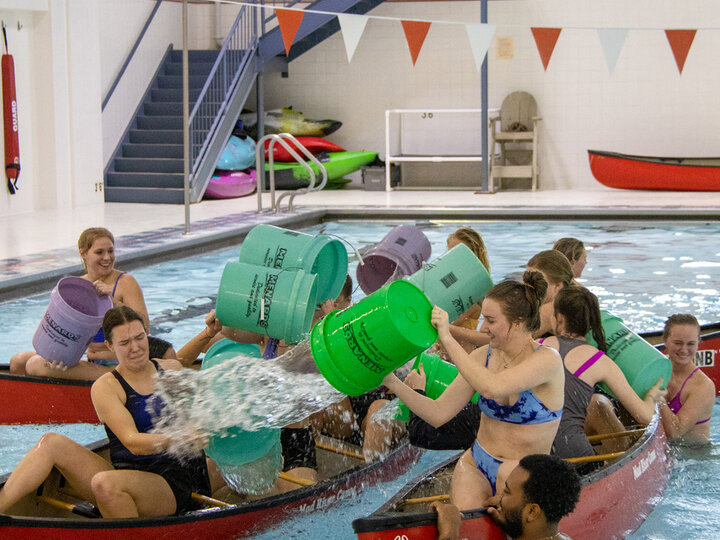 Two groups of people in canoes inside a swimming pool splash each other with buckets of water.