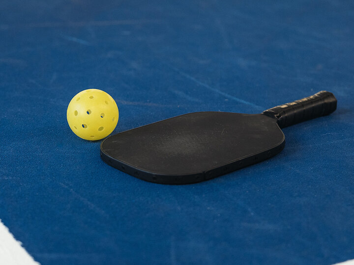 A yellow pickleball and a black paddle lie on a blue pickleball court.