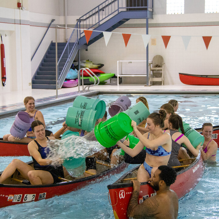 Two groups of people in canoes inside a swimming pool splash each other with buckets of water.