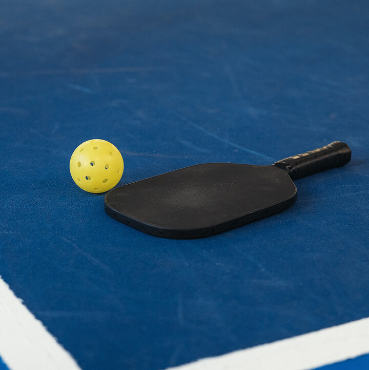 A yellow pickleball and a black paddle lie on a blue pickleball court.